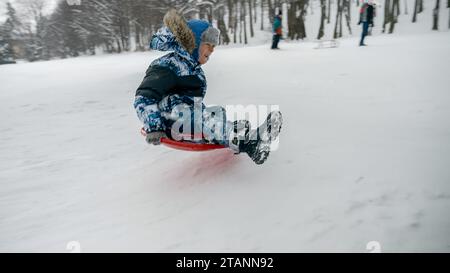 Junge lacht und lächelt, während er einen schneebedeckten Hügel hinunterfährt und auf seinem Plastikschlitten auf Trampolin springt. Winterferien und Kinder, die uns spielen Stockfoto
