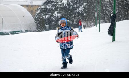 Ein glückliches Kind rennt mit Schlitten in der Hand durch den Schnee, um den Gipfel des Berges für eine aufregende Fahrt hinunter zu erreichen Stockfoto