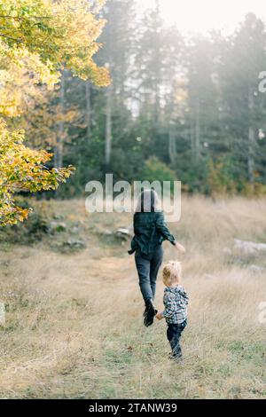 Das kleine Mädchen rennt ihrer Mutter über den Rasen in den Wald. Rückansicht Stockfoto