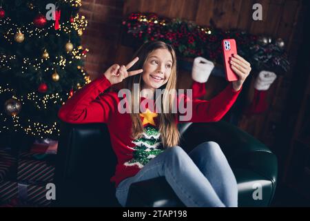 Porträt eines freundlichen, entzückenden Teenagers mit blondem Haar gekleidetem roten Pullover, der Selfie auf dem Smartphone macht, zeigt das V-Zeichen-Symbol zu Hause drinnen Stockfoto