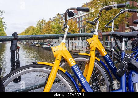 OV-fiets Leihfahrrad von NS, der niederländischen Eisenbahngesellschaft. Gelbes und blaues Fahrrad parkt auf der Brücke des Amsterdamer Kanals. Nahaufnahme. Amsterdam, Niederlande - 23. Oktober 2023 Stockfoto