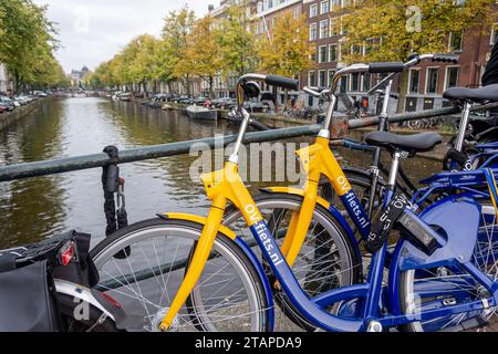 OV-fiets Leihfahrrad von NS, der niederländischen Eisenbahngesellschaft. Gelbes und blaues Fahrrad parkt auf der Brücke des Amsterdamer Kanals. Amsterdam, Niederlande - 23. Oktober 2023 Stockfoto