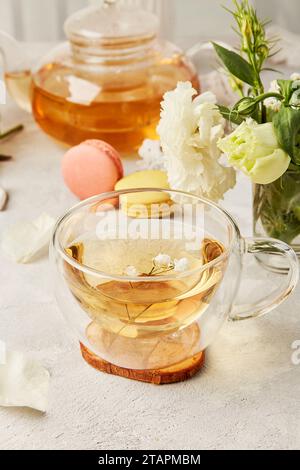 Kräuterteetasse und Makkarondesserts, ästhetische Teezeit im biophilen Innenraum unter weißen Blüten. Femininer ruhiger Lebensstil. Stockfoto