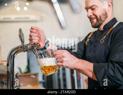 Ein lächelnder, stilvoller Bärenbarmann in schwarzer Uniform mit einer Schürze, der frisches Lagerbier in einen Glasbecher an der Bar zapft. Erfolgreiche Leute, Bierkonzern Stockfoto