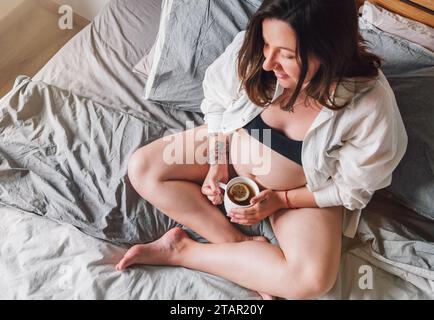 Junge schwangere Frau, die morgens mit gekreuzten Beinen auf dem Bett sitzt, mit einer Tasse Tee im Schlafzimmer. Frauengesundheit, glückliche Schwangerschaft und ca. Stockfoto