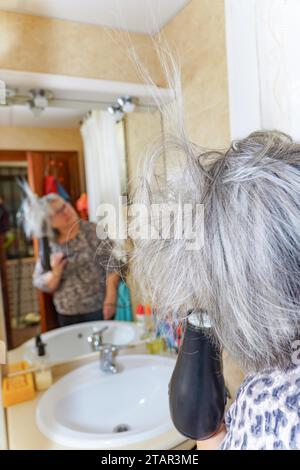 Reife weißhaarige Frau, die ihr Haar mit einem Handtrockner vor einem Spiegel trocknet Stockfoto