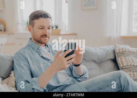 Fokussierter Mann, der ein Smartphone verwendet, SMS schreibt oder soziale Medien durchsucht, während er auf einem Sofa sitzt Stockfoto