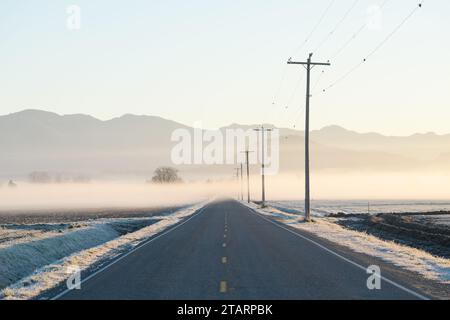 Dünne Nebelschicht über der Skagit Valley Road bei Sonnenaufgang mit Telefonmasten und Leitungen Stockfoto