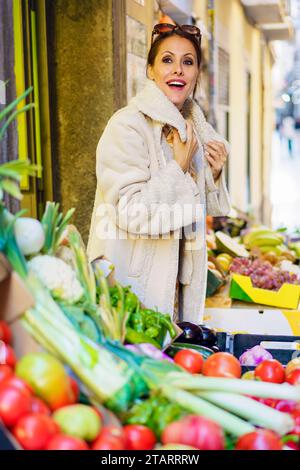 Lächelnde Frau in warmem Fell, die neben Obst und Gemüse steht Stockfoto