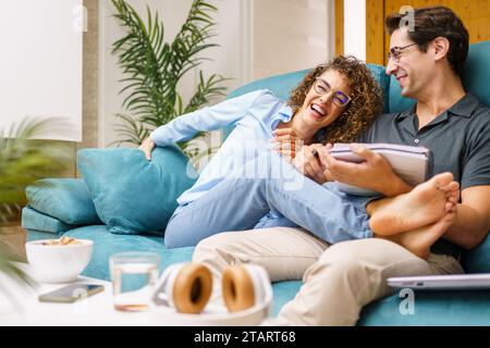 Glückliches Paar mit Notizbuch, das zu Hause auf dem Sofa sitzt und fröhlich lacht Stockfoto