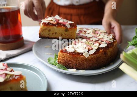 Frau, die ein Stück frisch gebackenen Rhabarberkuchen mit Mandelflocken am weißen Marmortisch nimmt, Nahaufnahme Stockfoto