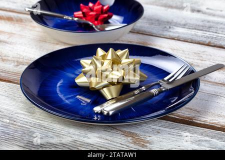 Nahansicht eines Feiertags-Esstisch mit dunkelblauem Hauptgericht und Schüssel mit Weihnachtsschmuck oder Silvester-Schleife plus Besteck auf rustikalem W Stockfoto