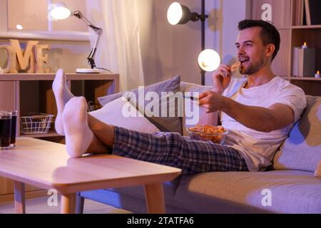 Junger Mann mit Kartoffelchips, der abends zu Hause fernsieht Stockfoto