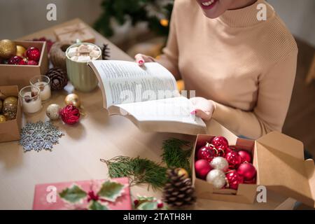 Junge asiatische Frau im Pullover, die ein Buch mit Glück liest und heiße Schokolade mit Marshmallow trinkt, nachdem sie Dekorationen zur Feier christi verziert hat Stockfoto