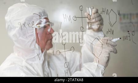 Ein älterer kaukasischer Chemiker im Schutzanzug schaut auf ein Reagenzglas und schreibt Formeln auf eine Glaswand. Stockfoto