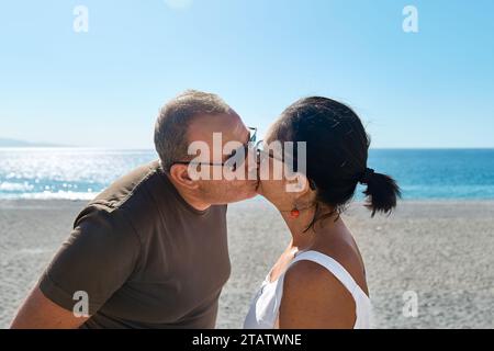 Glückliches Paar mittleren Alters, das Zeit zusammen verbringt und einen romantischen Moment am Strand genießt. Reifer lächelnder Mann und Frau, die sich am Meer umarmen und küssen. Stockfoto