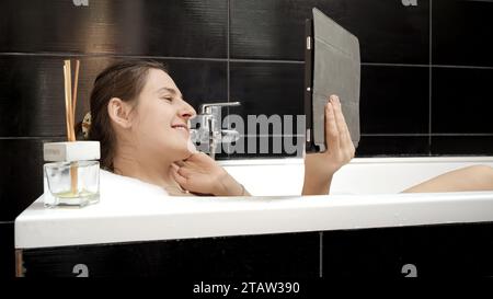 Frau, die ein Bad nimmt, während sie mit Freunden und Familie über einen Videoanruf auf ihrem Tablet in Verbindung bleibt. Authentizität und Förderung der Bedeutung Stockfoto