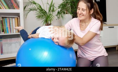 Porträt einer glücklichen lächelnden Mutter, die ihren Sohn hält, während sie ihn hält und sich auf dem Fitball ausdehnt. Konzept der Kinderbetreuung, Kindersport und Yoga Stockfoto