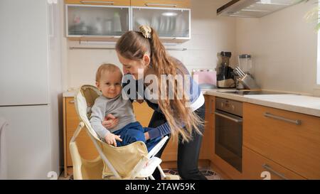 Die junge, fürsorgliche Mutter sitzt ihr Baby in einem Hochstuhl in der Küche, bevor sie frühstückt. Erziehungskonzept, gesunde Ernährung und Babypflege. Stockfoto