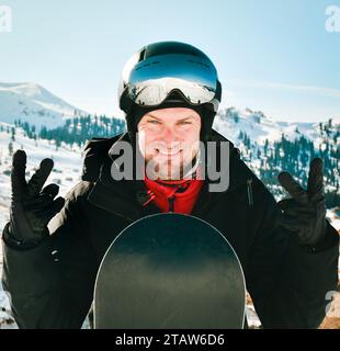 Kaukasischer Snowboarder mit Skibrille und farbenfroher Skiausrüstung, mit Daumen auf der Piste Stockfoto