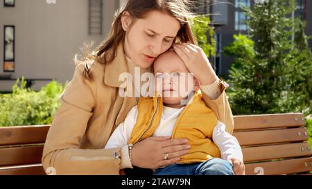 Kleiner kleiner Junge, der im Park auf den Beinen der Mutter sitzt und weint. Stockfoto