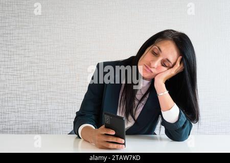 Junge, weiße, brünette Geschäftsfrau, Direktorin im Büro, sitzt am Schreibtisch, benutzt Telefon Scroll Nachrichten, soziale Medien gelangweilt am Arbeitsdirektor lustig. Unnötiges EM Stockfoto