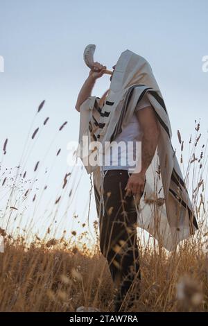 Jüdischer Mann in einem traditionellen Tallit-Gebetschal, der das Horn des Widders in Schofar bläst, auf dem Feld vor dem Sonnenaufgangshimmel auf Rosh HaShana und Yom Kippurim Stockfoto