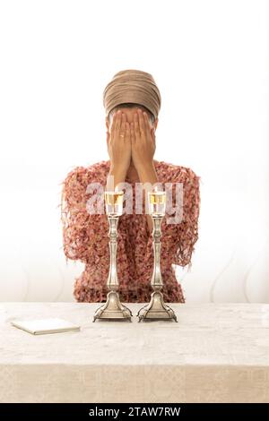 Jüdische Frau betet über zündete Shabbat-Kerzen und bedeckte ihr Gesicht mit ihren Händen. In der Nähe liegt ein religiöses Gebetsbuch. Stockfoto