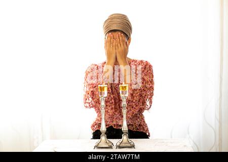 Jüdische Frau betet über zündete Shabbat-Kerzen und bedeckte ihr Gesicht mit ihren Händen. In der Nähe liegt ein religiöses Gebetsbuch. Stockfoto