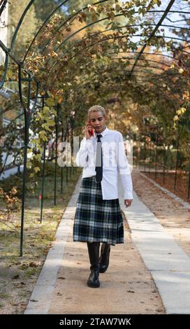 Ein junger Mann, der im Park läuft und einen Kilt trägt, der am Handy spricht. Stockfoto