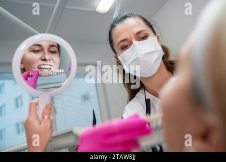 Junge Frau sitzt im Stomatologie-Klinikstuhl und lächelt im Spiegel, zeigt ihre Zähne und wählt Zahnfarben mit Farbtabellen. Gesundheit, Mund Stockfoto