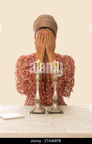 Jüdische Frau betet über zündete Shabbat-Kerzen und bedeckte ihr Gesicht mit ihren Händen. In der Nähe liegt ein religiöses Gebetsbuch. Stockfoto