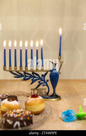 Menora (Chanukkia) mit 8 brennenden Kerzen für jüdischen Hanukka-Urlaub auf dem Tisch zu Hause. Dreidel und Donuts auf der Seite Stockfoto