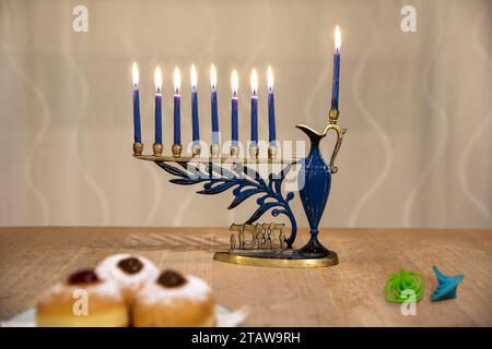 Menora (Chanukkia) mit 8 brennenden Kerzen für jüdischen Hanukka-Urlaub auf dem Tisch zu Hause. Dreidel und Donuts auf der Seite Stockfoto