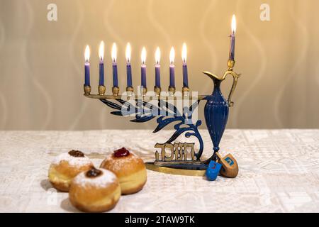 Menora (Chanukkia) mit 8 brennenden Kerzen für jüdischen Hanukka-Urlaub auf dem Tisch zu Hause. Dreidel und Donuts auf der Seite Stockfoto