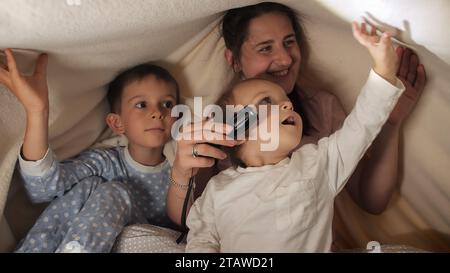 Zwei Jungs und Mutter im Pyjama spielen mit Taschenlampe, während sie sich nachts unter der Decke verstecken. Familie, die Zeit zusammen hat, Eltern, glückliche Kindheit und Stockfoto