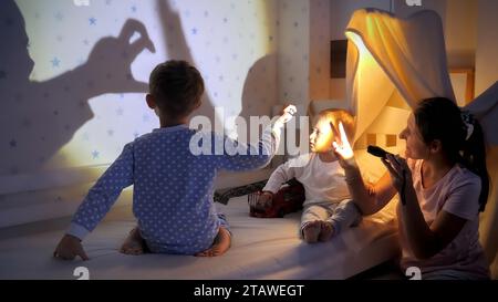 Die junge Mutter spielt mit Taschenlampe und ihre beiden Söhne zeigen ihnen Schatten an der Wand. Stockfoto