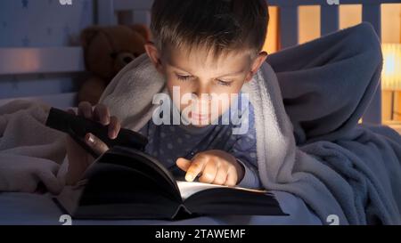 Porträt eines Jungen mit Fackel, der im Bett liegt und Geschichtenbuch liest. Kindererziehung, Entwicklung, Geheimhaltung, Privatsphäre, Bücher lesen Stockfoto