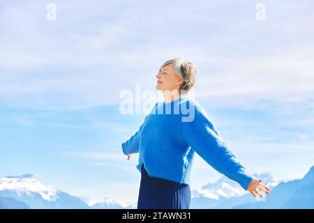 Außenporträt der glücklichen mittleren 55-60-jährigen Frau mit weit geöffneten Armen, posiert auf blauem Himmel und Berghintergrund Stockfoto