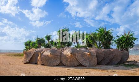 Strohschirm am Strand. Sonnenschirm aus Bambus-Strohhalm, entfernt und auf seinem Stand aufgelegt, steht in einer Reihe an einem Sandstrand mit blauem Himmel. Stockfoto