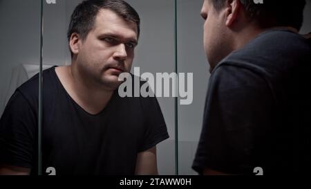 Ein verärgerter Mann, der sich auf das Waschbecken lehnt und im Spiegel ins Badezimmer blickt. Vorstellung von Depression, Suizid, Stress, Geisteskrankheiten, Einsamkeit und Frustration Stockfoto