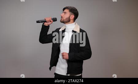 Ein junger Mann, der ein Mikrofon hält und vor grauem Hintergrund singt. Stockfoto