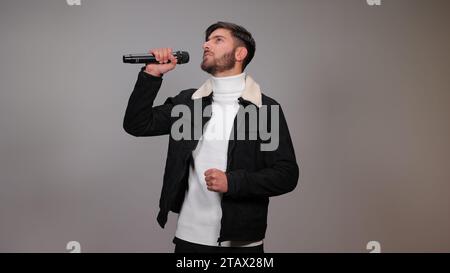 Ein junger Mann, der ein Mikrofon hält und vor grauem Hintergrund singt. Stockfoto