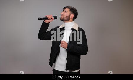 Ein junger Mann, der ein Mikrofon hält und vor grauem Hintergrund singt. Stockfoto