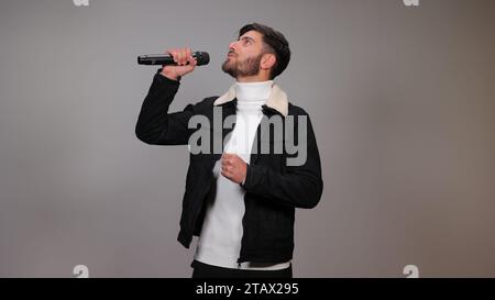 Ein junger Mann, der ein Mikrofon hält und vor grauem Hintergrund singt. Stockfoto