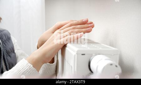 Nahaufnahme einer jungen Frau im Pullover, die ihre Hände an der Heizung im kalten Zuhause wärmt. Stockfoto