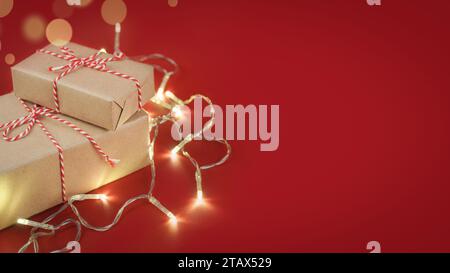 Weihnachtsgeschenkboxen in Bastelpapier mit gestreiftem rot-weißem Backgarn mit Lichtern von Girlande auf rotem Hintergrund mit Kopierraum. Flach-La Stockfoto