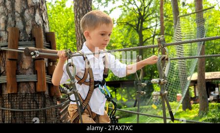 Porträt eines niedlichen kleinen Jungen, der auf der Seilbrücke spaziert, während er Spaß im Pfadfindercamp hat. Aktive Kindheit, gesunder Lebensstil, Kinder spielen draußen, Chi Stockfoto