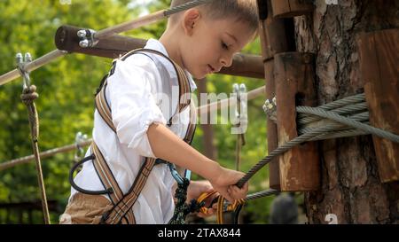 Kleiner Junge klemmt Sicherheitsseile am Baum, während er im Seilpark klettert. Aktive Kindheit, gesunder Lebensstil, Kinder spielen draußen, c Stockfoto