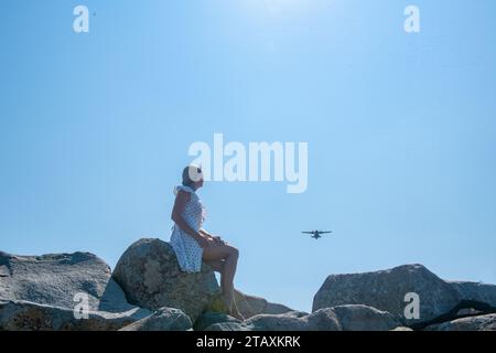 Ein Mädchen in einem weißen Kleid sitzt auf grauen Felsen und schaut sich ein Flugzeug an Stockfoto